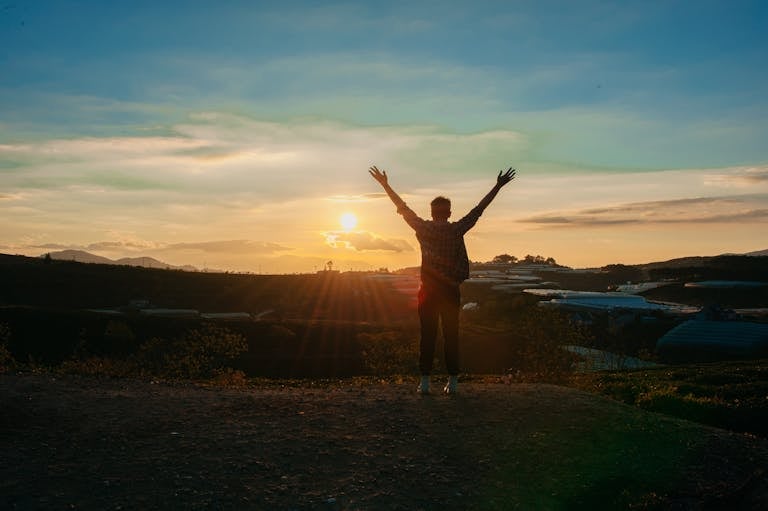 A man raising both hands in an outdoor setting, looking at a sunset, symbolizing gratitude, freedom, and new beginnings.